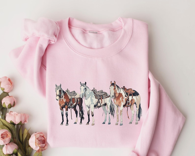 May include: Pink sweatshirt with a graphic of four horses wearing saddles. The horses are brown, white, and black.