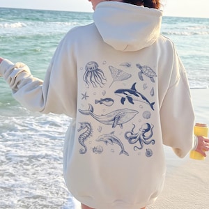 May include: Cream-colored hoodie featuring a blue illustration of various marine animals, including a whale, dolphin, and octopus. The design is on the back of the hoodie. The hoodie has a hood and long sleeves.