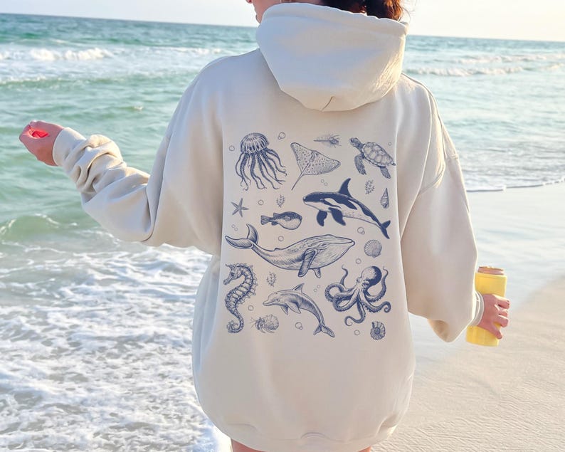 May include: Cream-colored hoodie featuring a blue illustration of various marine animals, including a whale, dolphin, and octopus. The design is on the back of the hoodie. The hoodie has a hood and long sleeves.