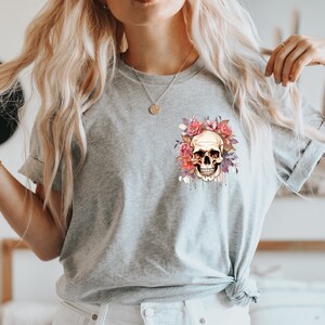 May include: A light gray t-shirt featuring a skull design surrounded by pink and purple flowers. The skull has a detailed, realistic appearance with dripping accents. The design is centered on the chest.