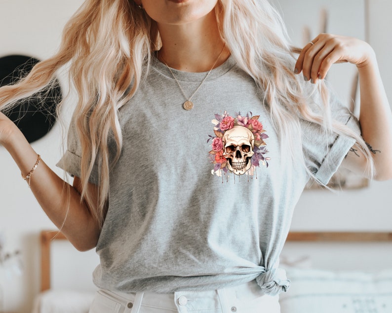 May include: A light gray t-shirt featuring a skull design surrounded by pink and purple flowers. The skull has a detailed, realistic appearance with dripping accents. The design is centered on the chest.