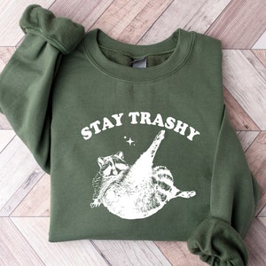 Op de afbeelding: Een groen sweatshirt met een witte afbeelding van een wasbeer en de tekst "STAY TRASHY".
