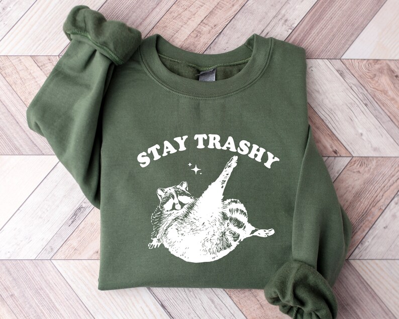 Op de afbeelding: Een groen sweatshirt met een witte afbeelding van een wasbeer en de tekst "STAY TRASHY".