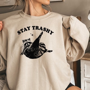 Op de afbeelding: Een beige sweatshirt met een zwarte afbeelding van een wasbeer met de tekst "STAY TRASHY" erboven.