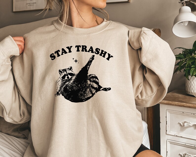 Op de afbeelding: Een beige sweatshirt met een zwarte afbeelding van een wasbeer met de tekst "STAY TRASHY" erboven.