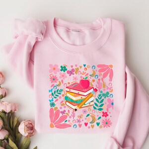 May include: Pink sweatshirt with a colorful graphic of a stack of books, an apple, and a pencil surrounded by flowers and hearts. The text "Read. Learn. Grow." is printed on the sweatshirt.
