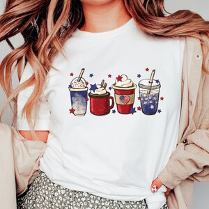 May include: White t-shirt featuring a graphic of four festive beverages. Each drink is decorated with whipped cream, straws, and patriotic red, white, and blue star accents. The drinks are in clear cups and a red mug.