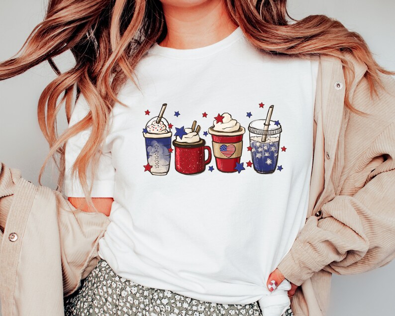 May include: White t-shirt featuring a graphic of four festive beverages. Each drink is decorated with whipped cream, straws, and patriotic red, white, and blue star accents. The drinks are in clear cups and a red mug.