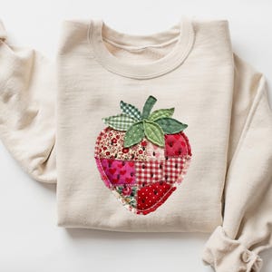 May include: A cream-coloured crewneck sweatshirt featuring a large, central strawberry appliqu&eacute;. The strawberry is constructed from various red and pink fabric patches with floral, heart, and gingham patterns, accented by green fabric leaves.