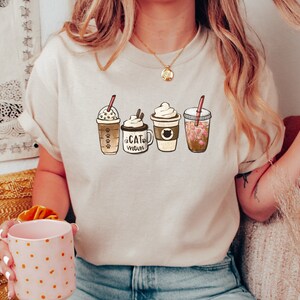 以下が含まれることがあります： 4種類の異なるコーヒー飲料のグラフィックが描かれたベージュのTシャツ。シャツのテキストは「Cat Mom」です。