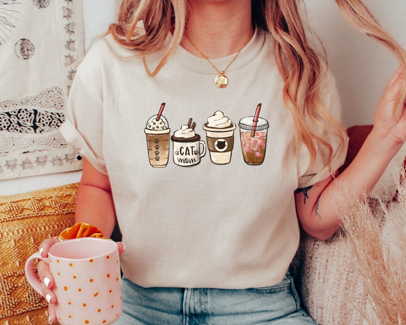 以下が含まれることがあります： 4種類の異なるコーヒー飲料のグラフィックが描かれたベージュのTシャツ。シャツのテキストは「Cat Mom」です。