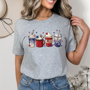 May include: A heather gray t-shirt featuring a graphic of four festive beverages, each adorned with patriotic red, white, and blue decorations. The drinks include whipped cream, stars, and a flag design, perfect for celebrating a holiday.