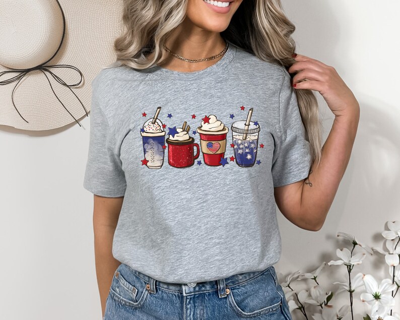 May include: A heather gray t-shirt featuring a graphic of four festive beverages, each adorned with patriotic red, white, and blue decorations. The drinks include whipped cream, stars, and a flag design, perfect for celebrating a holiday.