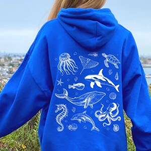 May include: Royal blue hoodie featuring a white illustration of various sea creatures, including a whale, dolphin, and octopus. The design also includes a jellyfish, stingray, and sea turtle. The hoodie is made of a soft material and has a hood.