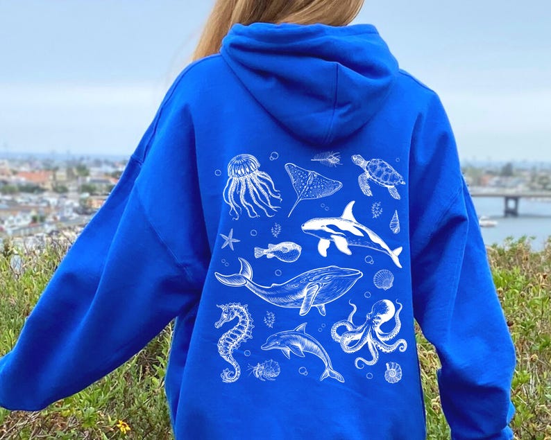 May include: Royal blue hoodie featuring a white illustration of various sea creatures, including a whale, dolphin, and octopus. The design also includes a jellyfish, stingray, and sea turtle. The hoodie is made of a soft material and has a hood.