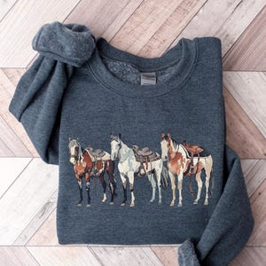 May include: A dark gray sweatshirt with a graphic of four horses wearing saddles. The horses are brown, white, and black with brown, white, and black saddles.