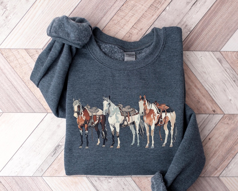 May include: A dark gray sweatshirt with a graphic of four horses wearing saddles. The horses are brown, white, and black with brown, white, and black saddles.