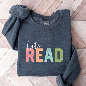 May include: A dark gray sweatshirt with the text "Let's READ" in colorful, distressed letters. The letters are in shades of pink, orange, blue, and green.