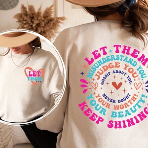 May include: White sweatshirt with a pink, blue, and white graphic design that says "Let them misunderstand you, judge you, gossip about you. Never doubt your worth! Your beauty! Keep shining!"