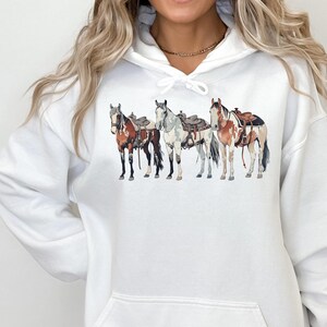 May include: White hoodie with a graphic of four horses wearing saddles. The horses are brown, white, and black.