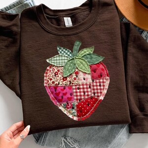 May include: A dark brown long-sleeve sweatshirt featuring a large, central strawberry-shaped appliqu&eacute;. The strawberry is constructed from various fabric patches in shades of red, pink, and white, featuring floral, heart, and gingham patterns.