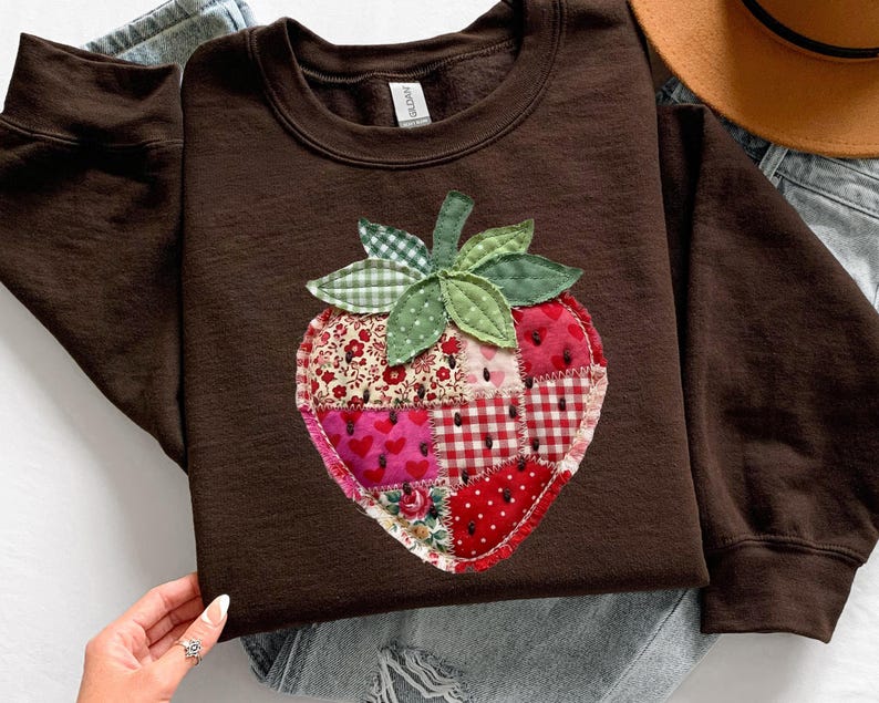 May include: A dark brown long-sleeve sweatshirt featuring a large, central strawberry-shaped appliqu&eacute;. The strawberry is constructed from various fabric patches in shades of red, pink, and white, featuring floral, heart, and gingham patterns.
