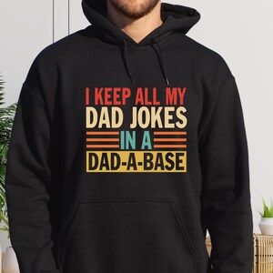 May include: Black hooded sweatshirt with a vintage-style graphic that reads "I keep all my dad jokes in a dad-a-base".