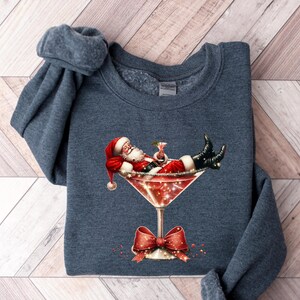 May include: A dark blue sweatshirt with a festive design featuring Santa Claus relaxing in a martini glass. The design includes red, white, and green colors, and glitter accents.