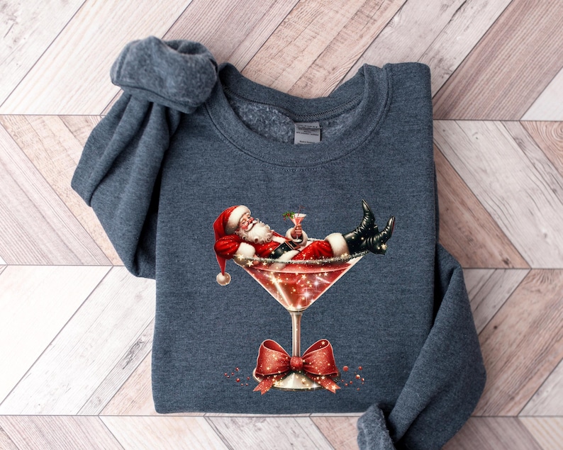 May include: A dark blue sweatshirt with a festive design featuring Santa Claus relaxing in a martini glass. The design includes red, white, and green colors, and glitter accents.