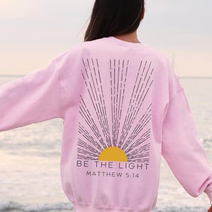 May include: Pink sweatshirt with a black and yellow sun graphic and the text "BE THE LIGHT" and "MATTHEW 5:14" printed on the back.