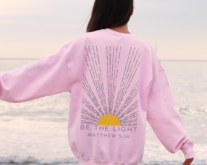 May include: Pink sweatshirt with a black and yellow sun graphic and the text "BE THE LIGHT" and "MATTHEW 5:14" printed on the back.