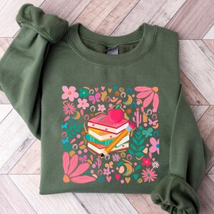 May include: A green sweatshirt with a colorful graphic print of a stack of books, an apple, and a pencil surrounded by flowers and butterflies.