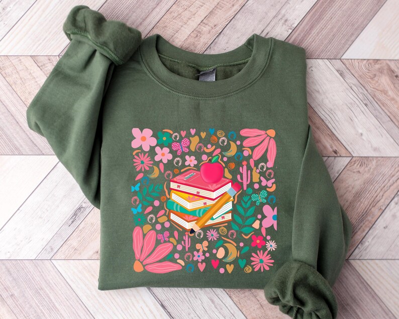 May include: A green sweatshirt with a colorful graphic print of a stack of books, an apple, and a pencil surrounded by flowers and butterflies.