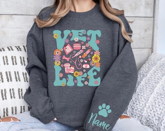 May include: A dark gray sweatshirt with the text "VET LIFE" in a retro font surrounded by colorful flowers and veterinary symbols. The text "Name" is on the sleeve with a paw print.