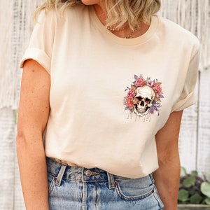 May include: A cream-colored t-shirt featuring a watercolor design of a skull surrounded by pink and purple roses. The skull has a detailed, realistic appearance. The shirt has rolled-up sleeves and is paired with blue jeans.