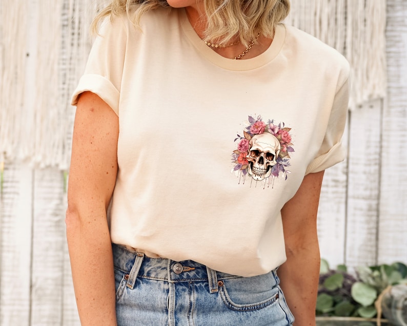 May include: A cream-colored t-shirt featuring a watercolor design of a skull surrounded by pink and purple roses. The skull has a detailed, realistic appearance. The shirt has rolled-up sleeves and is paired with blue jeans.
