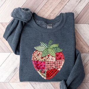 May include: A dark heather grey crewneck sweatshirt featuring a large, central strawberry-shaped appliqu&eacute;. The strawberry is constructed from various patchwork fabric squares in shades of red, pink, and white, decorated with small black seed details.