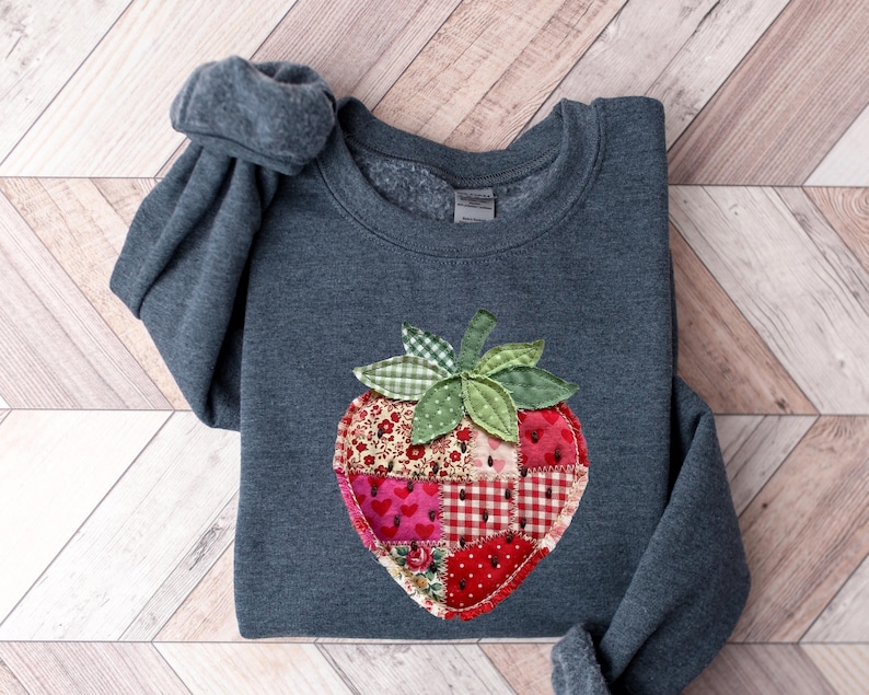 May include: A dark heather grey crewneck sweatshirt featuring a large, central strawberry-shaped appliqu&eacute;. The strawberry is constructed from various patchwork fabric squares in shades of red, pink, and white, decorated with small black seed details.