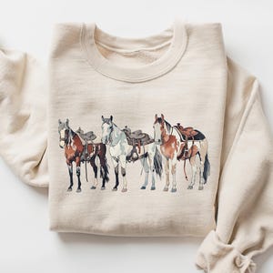 May include: A beige sweatshirt with a graphic print of four horses wearing saddles. The horses are brown, white, and black.