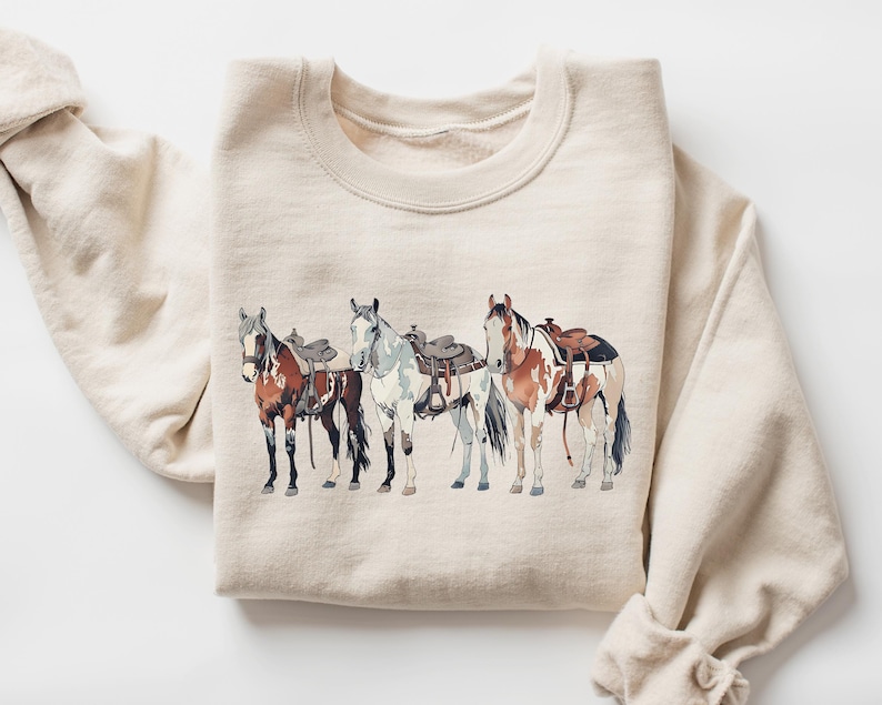 May include: A beige sweatshirt with a graphic print of four horses wearing saddles. The horses are brown, white, and black.