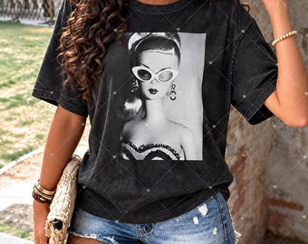 May include: A charcoal black, acid-wash t-shirt featuring a centered, black-and-white graphic print of a doll wearing cat-eye sunglasses and a strapless top. The shirt has short sleeves and a crew neckline, showcasing a vintage-inspired aesthetic.