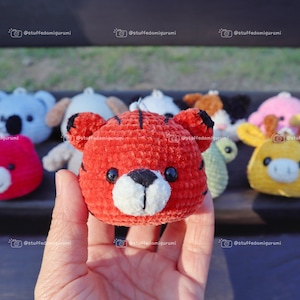 May include: A close-up of a red crocheted tiger plushie with white accents. The tiger is being held in a hand and there are other crocheted plushies in the background.