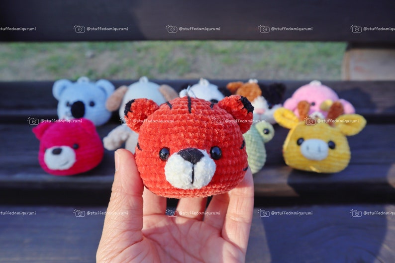 May include: A close-up of a red crocheted tiger plushie with white accents. The tiger is being held in a hand and there are other crocheted plushies in the background.