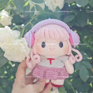 May include: A crocheted doll with pink hair, a pink and purple headband, and a pink and gray outfit. The doll is holding a pair of headphones and is smiling.