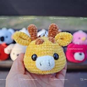 May include: A yellow crocheted giraffe plushie with brown ears and a white nose. The giraffe is being held in a hand. There are other crocheted plushies in the background.