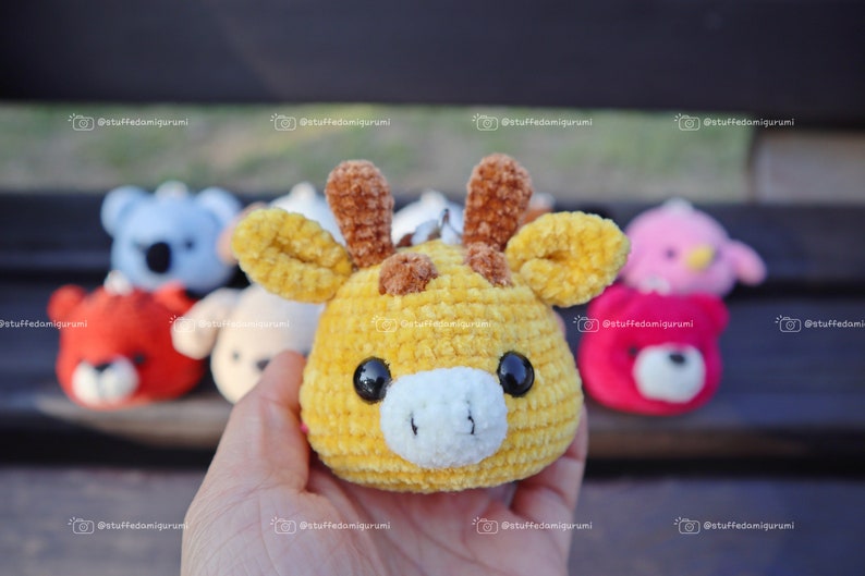 May include: A yellow crocheted giraffe plushie with brown ears and a white nose. The giraffe is being held in a hand. There are other crocheted plushies in the background.