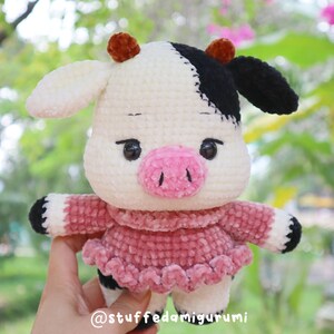 Peut inclure: Un jouet en peluche de vache crocheté portant une robe rose. Le jouet a des taches noires et blanches et des cornes brunes. Le jouet est tenu par une main. La légende sur l'image est "@stuffedamigurumi".