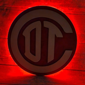 May include: A round, red and white logo with the letters "DT" and "C" in a stylized font. The logo is set against a red wooden background, with a bright red glow around the edges.