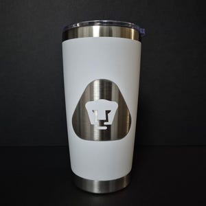 May include: A white stainless steel tumbler with a clear plastic lid. The exterior features a brushed silver triangular emblem centered on the front, displaying a white stylized puma head logo within the metallic shape of the shield.