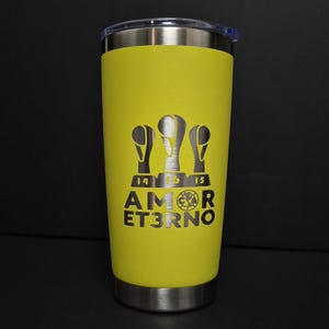May include: A bright yellow stainless steel tumbler with a clear lid. The front features a laser-engraved design of three trophy icons above the text AMOR ET3RNO, with the numbers 14, 16, and 15 positioned below the trophies.
